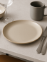 Matowy, beżowy ceramiczny talerz leży pośrodku jasnoszarego blatu. Minimalistyczna aranżacja stołu obejmuje szary kubek, srebrne sztućce oraz pusty kieliszek.