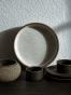 Duży ceramiczny talerz z pofalowanym, strukturalnym wzorem przypominającym piasek opiera się o białą, chropowatą ścianę. W mocnym świetle słonecznym otacza go dopasowany zestaw naczyń w ciemnobrązowym kolorze, w tym małe czarki, miska i spodki.