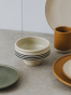 Kremowa ceramiczna miska na podwyższeniu z czarnymi, poziomymi paskami stoi na szarym blacie. Minimalistyczna aranżacja naczyń obejmuje talerze w kolorach ziemi oraz ochrowy kubek w tle.